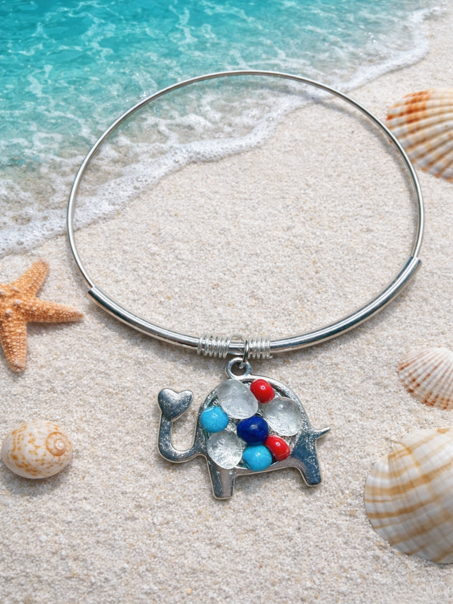 Silver bangle with an elephant charm featuring blue and red stones on a sandy beach with shells and water.
