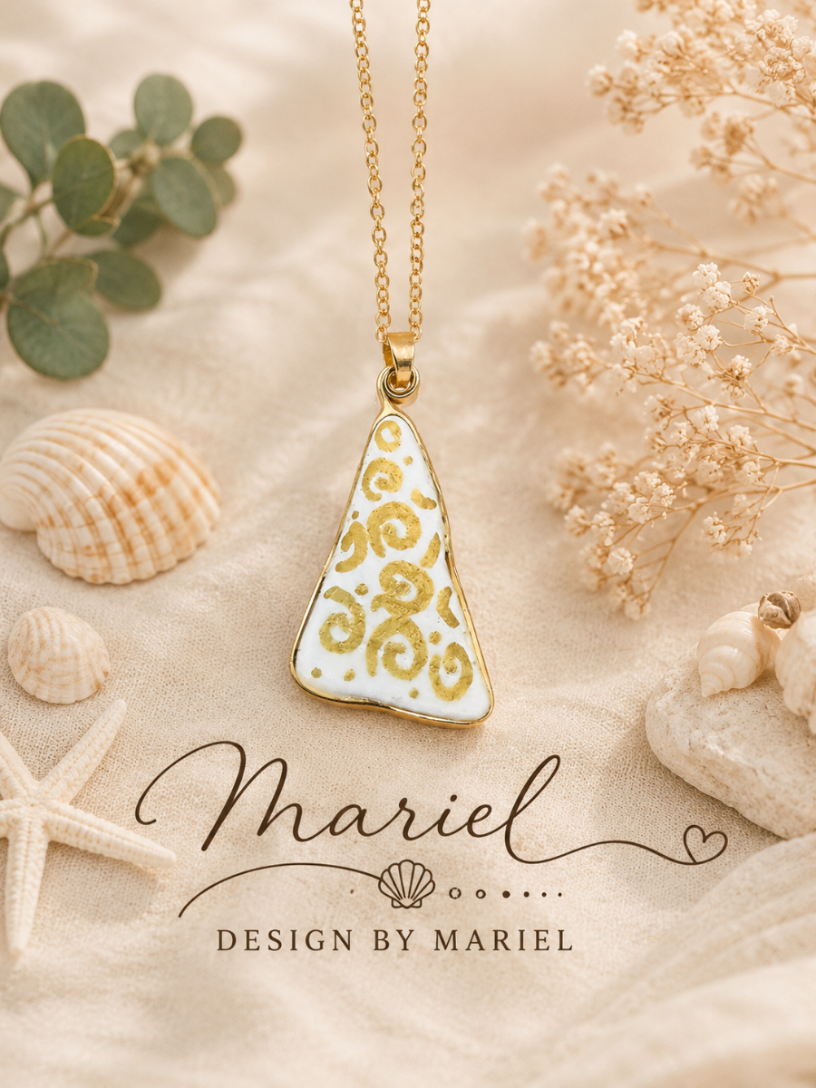 Gold necklace with a decorative pendant on a textured surface with shells and flowers, featuring 'Design by Mariel' branding.