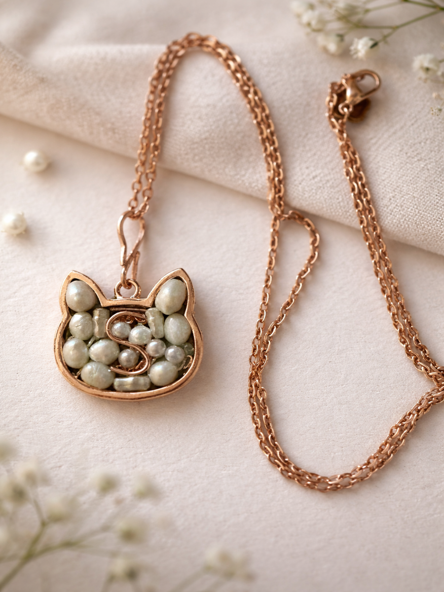 A rose gold cat-shaped pendant necklace with pale green and white faux pearls, displayed on a soft beige fabric background with small white flowers.