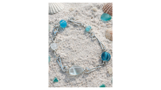 Handcrafted silver bracelet featuring sea glass beads in soft aqua and white tones, displayed on sand with shells for a coastal, beach-inspired look.