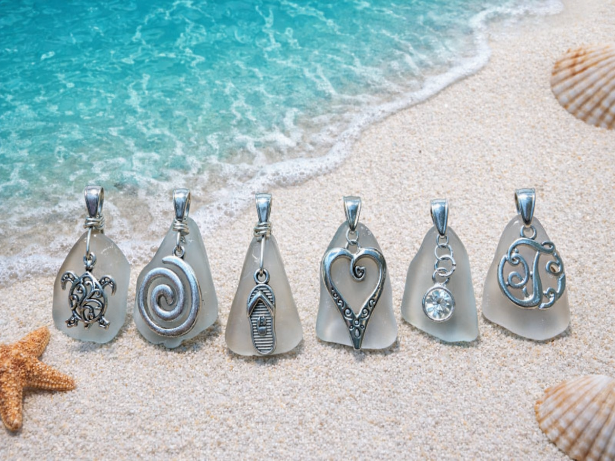 Sea glass pendants with various designs on a sandy beach with ocean waves.
