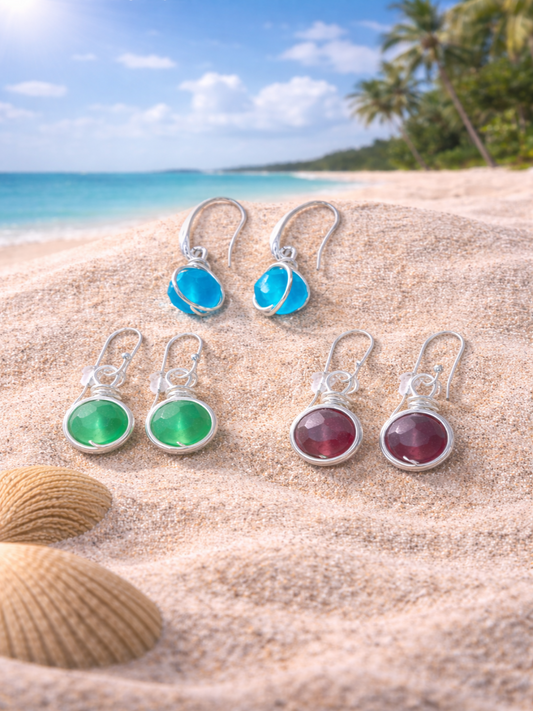 Set of colorful gemstone earrings on a sandy beach with ocean and palm trees in the background.