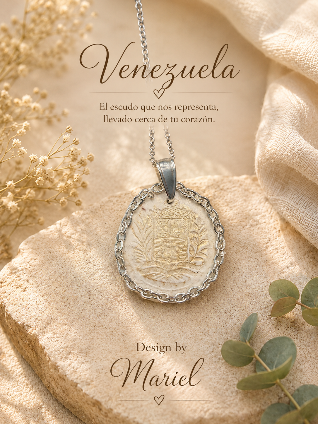 A silver pendant necklace with an oval engraved plaque featuring a Venezuelan coat of arms, styled on a stone slab with beige fabric, dried flowers, and green leaves. Spanish text reads 'Venezuela' and 'Design by Mariel'.