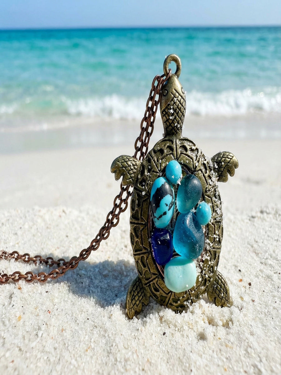 Tortoise-shaped necklace with blue and green stones on a sandy beach