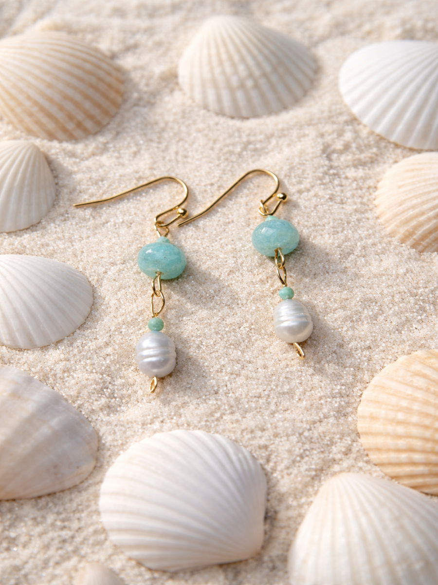 Gold earrings with turquoise and pearl beads on a sandy surface with seashells.