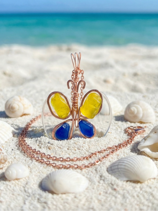 Butterfly-shaped necklace with blue and yellow wings on a sandy beach with seashells.