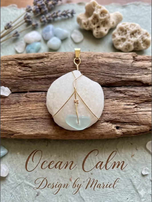 A handmade pendant necklace featuring a round, textured white stone with a small light blue bead at the bottom, wrapped in gold wire. The pendant is displayed on a piece of weathered wood with decorative stones and lavender sprigs in the background, with the text 'Ocean Calm Design by Mariel' written at the bottom.